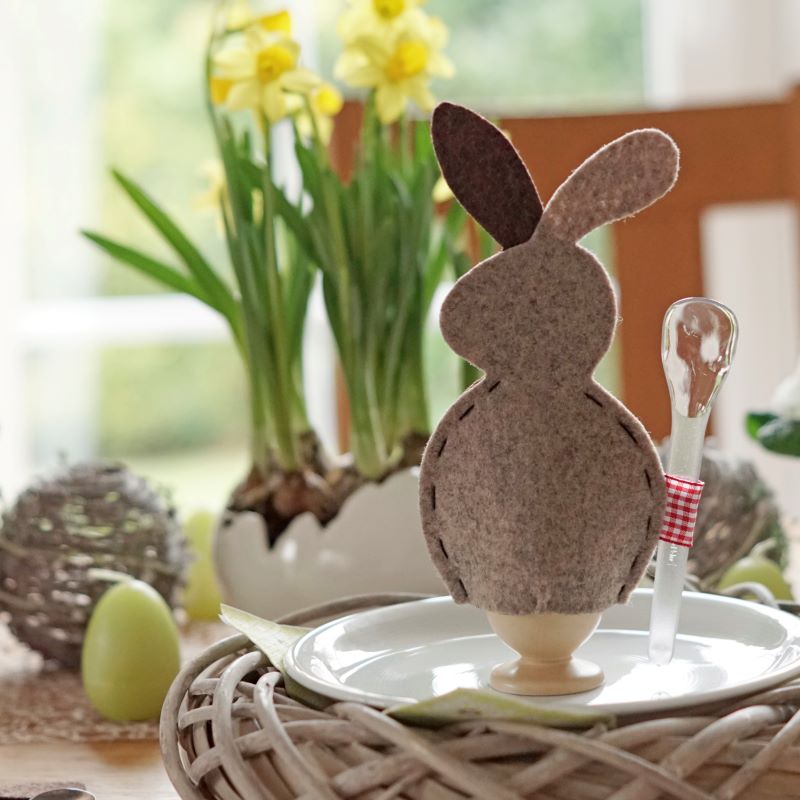 Hasen-Eierwärmer aus Filz auf gedeckten Tisch mit Osterglocken, Eierkerzen und Rattan-Kränzen